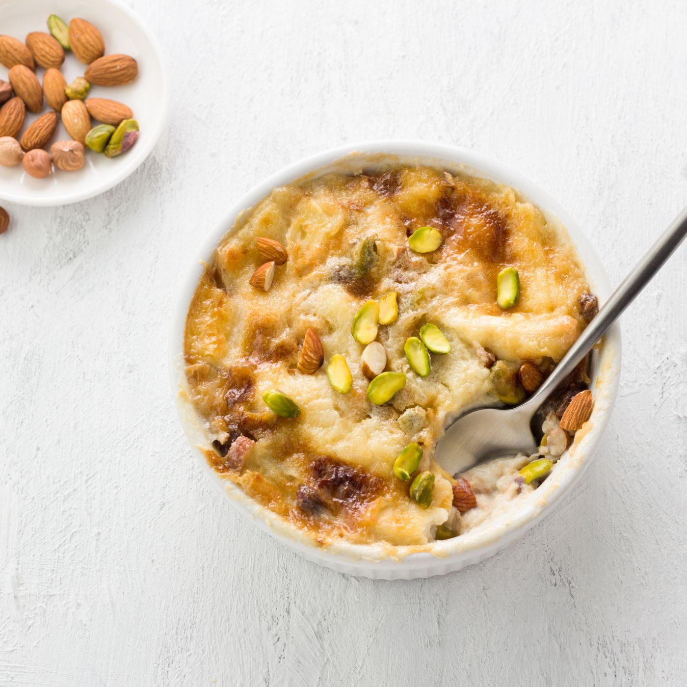 Traditional-Arabic-dessert-Umm-Ali-in-a-white-ceramic-form-decorated-with-nuts-on-a-light-gray-background-1490124906_2136x1408 square.jpg