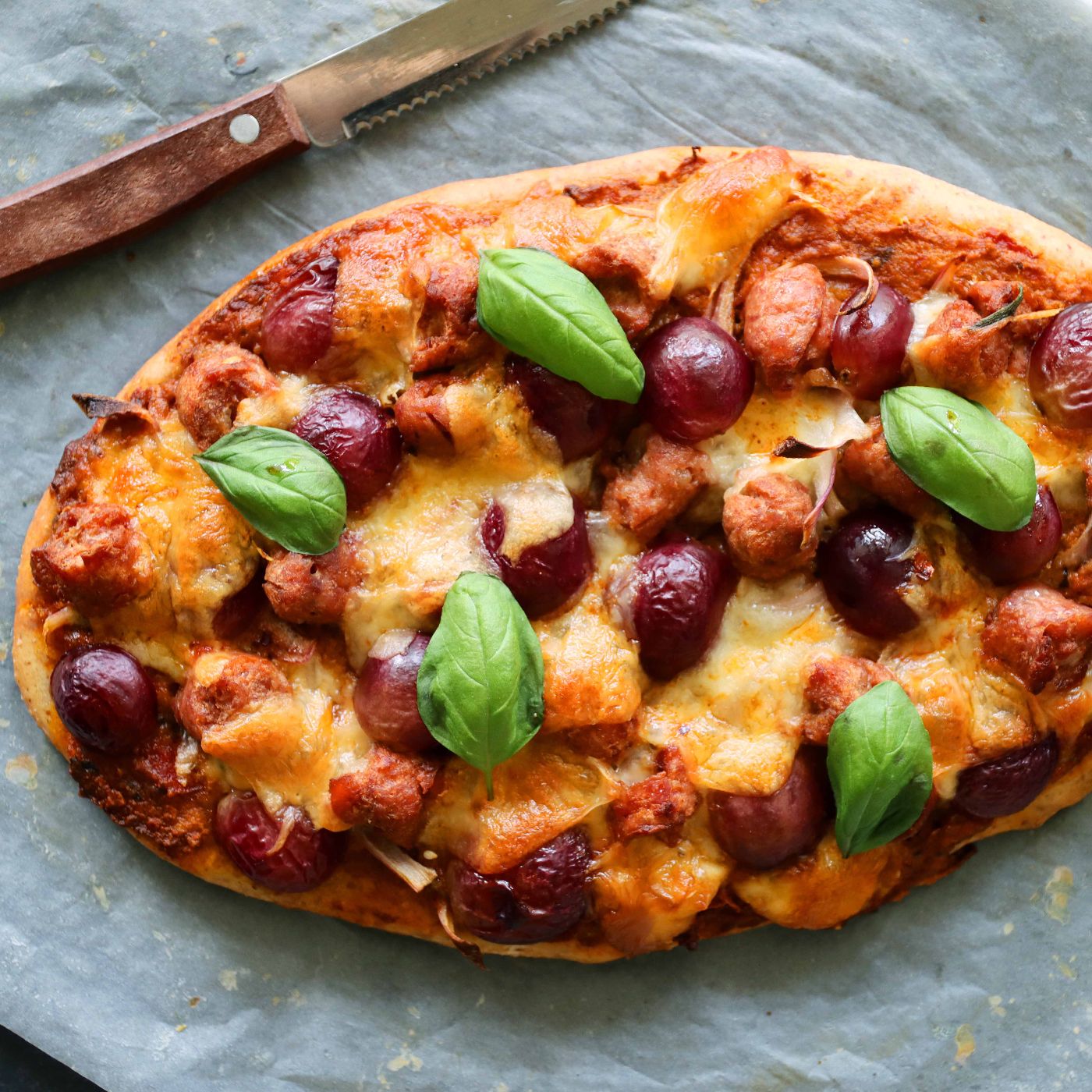 Image-of-freshly-baked-homemade-pizza-on-greaseproof-paper-with-knife-sausage-meatballs-grapes-mozzarella-cheese-basil-leaf-topping-rich-tomato-sauce-elevated-view-focus-on-foreground-1329673937_6960x4640_square.jpeg